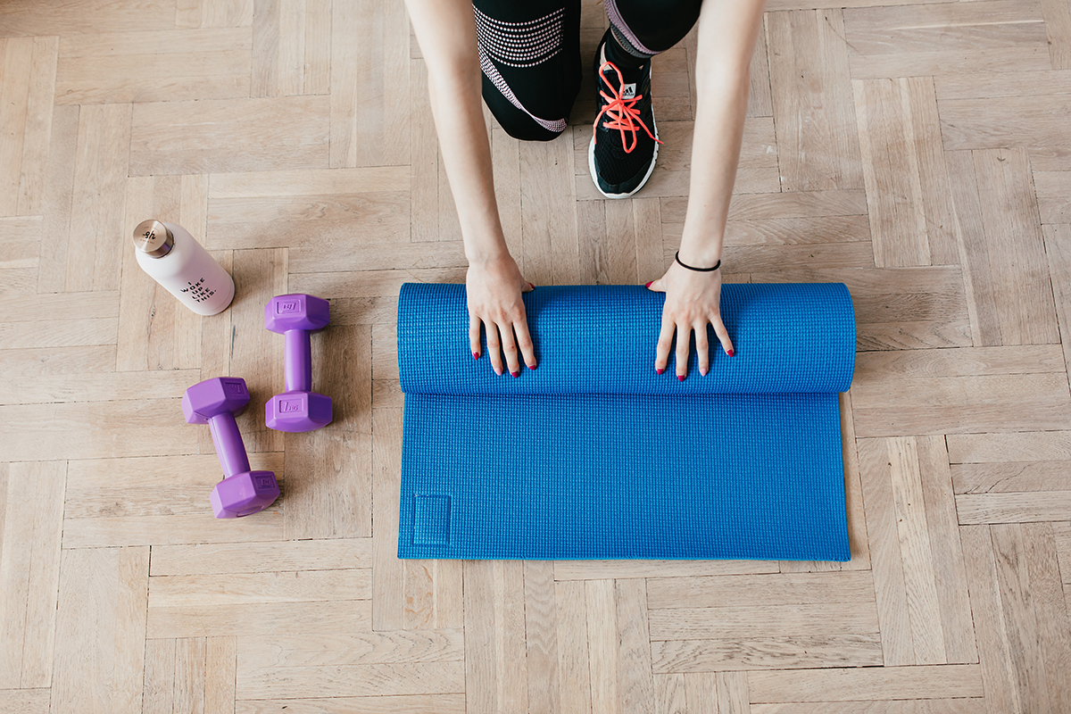 En dame ruller en blå yogamatte med ett par lilla treningsvekter ved siden av og en flaske.